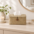 A rectangular woven straw tissue box cover with a slotted top and a small brown leather tab closure, photographed on a dressing table space.