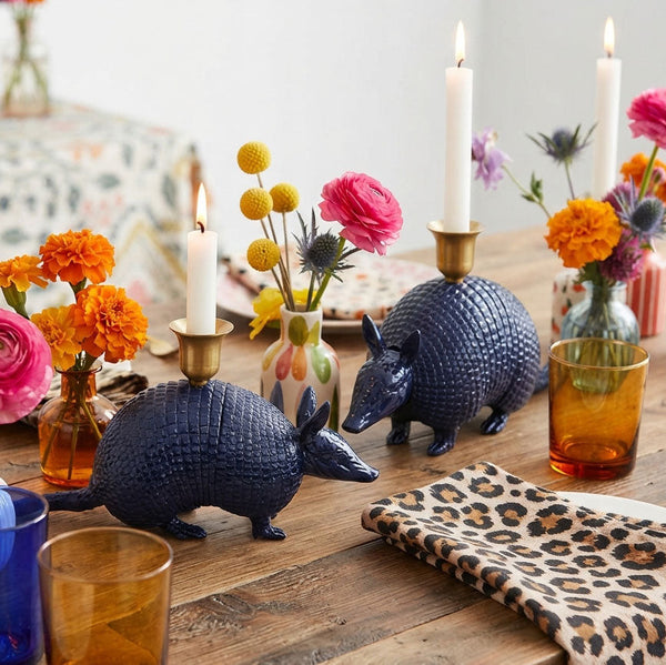 Two dark blue armadillo-shaped candle holders on a wooden table, styled with white taper candles, colorful flower vases, amber glasses, and a leopard-print napkin.