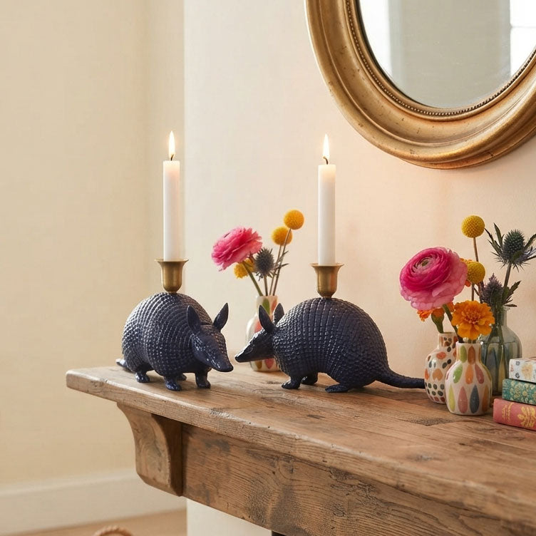 Two navy armadillo-shaped candle holders with brass candle sleeves and lit white taper candles, displayed on a rustic wooden mantel with flowers and a gold mirror in the background.